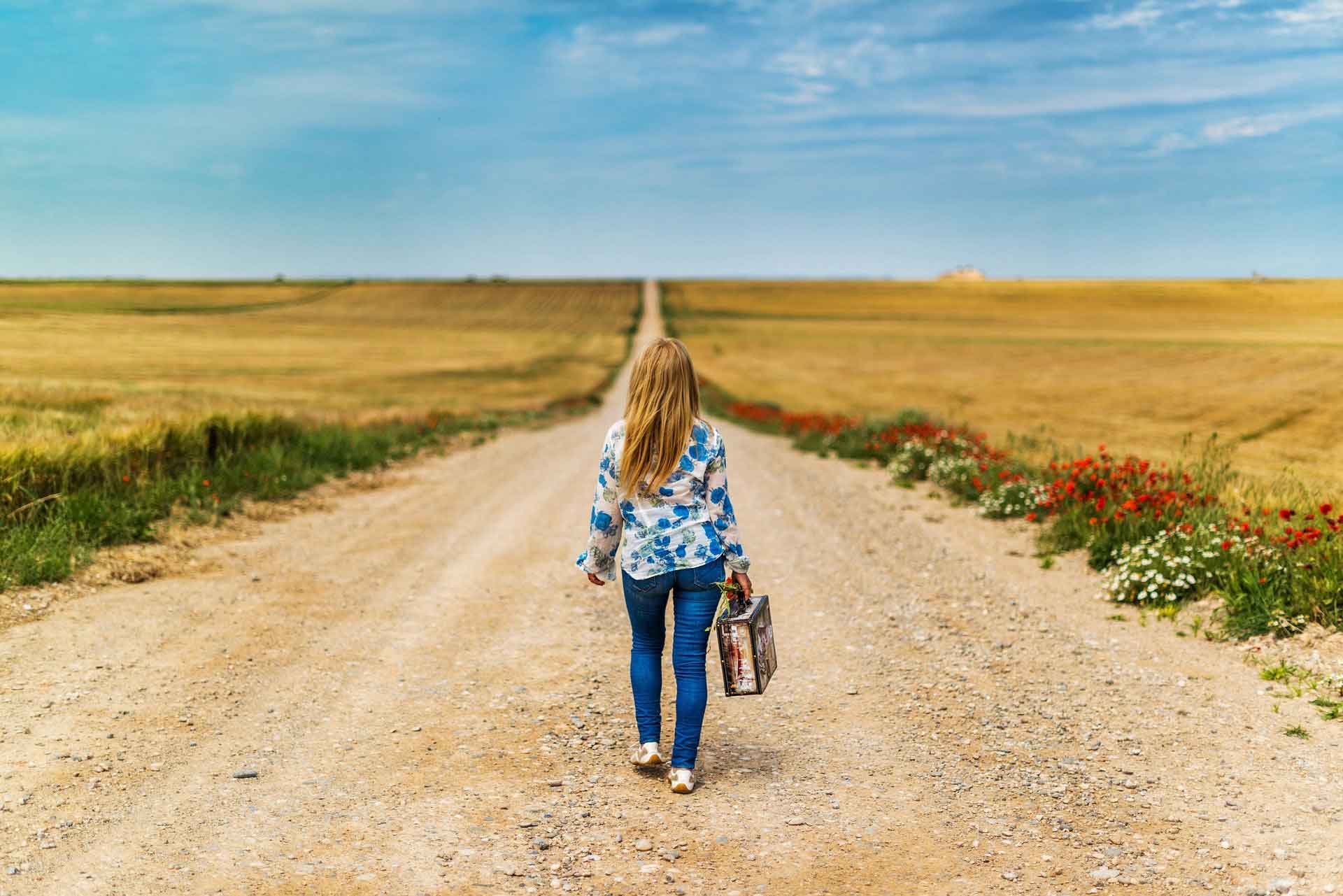 Nainen kävelee matkalaukkuk kädessä keskellä hiekkatietä, jota ympäröivät viljapellot.