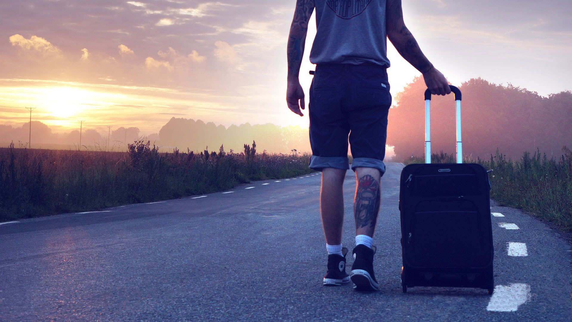 Shortsiasuinen mies kävelee maalaismaisemassa tietä pitkin ilta-auringon aikaan ja vetää matkalaukkua perässään.